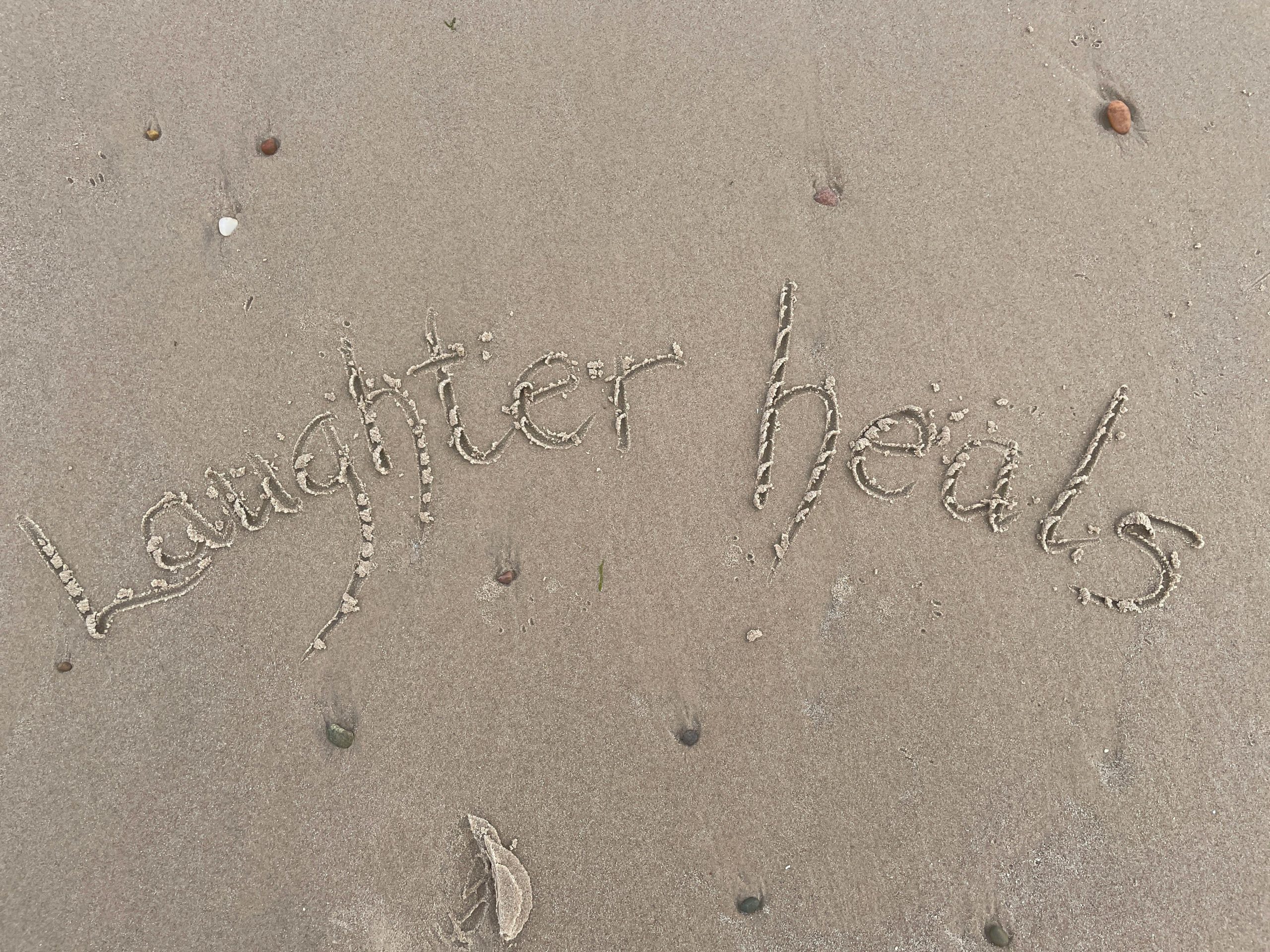 Inspirational 'Laughter Heals' written on a serene Scottish beach sand.